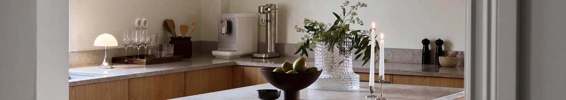 Modern kitchen counter with a mushroom lamp, wine glasses, Smeg water dispenser, vase with greenery, pears, and lit candles.
