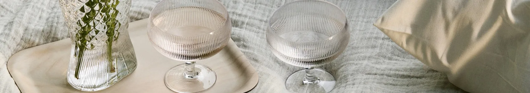 Two ribbed glass goblets and a vase with foliage on a tray and textured fabric.
