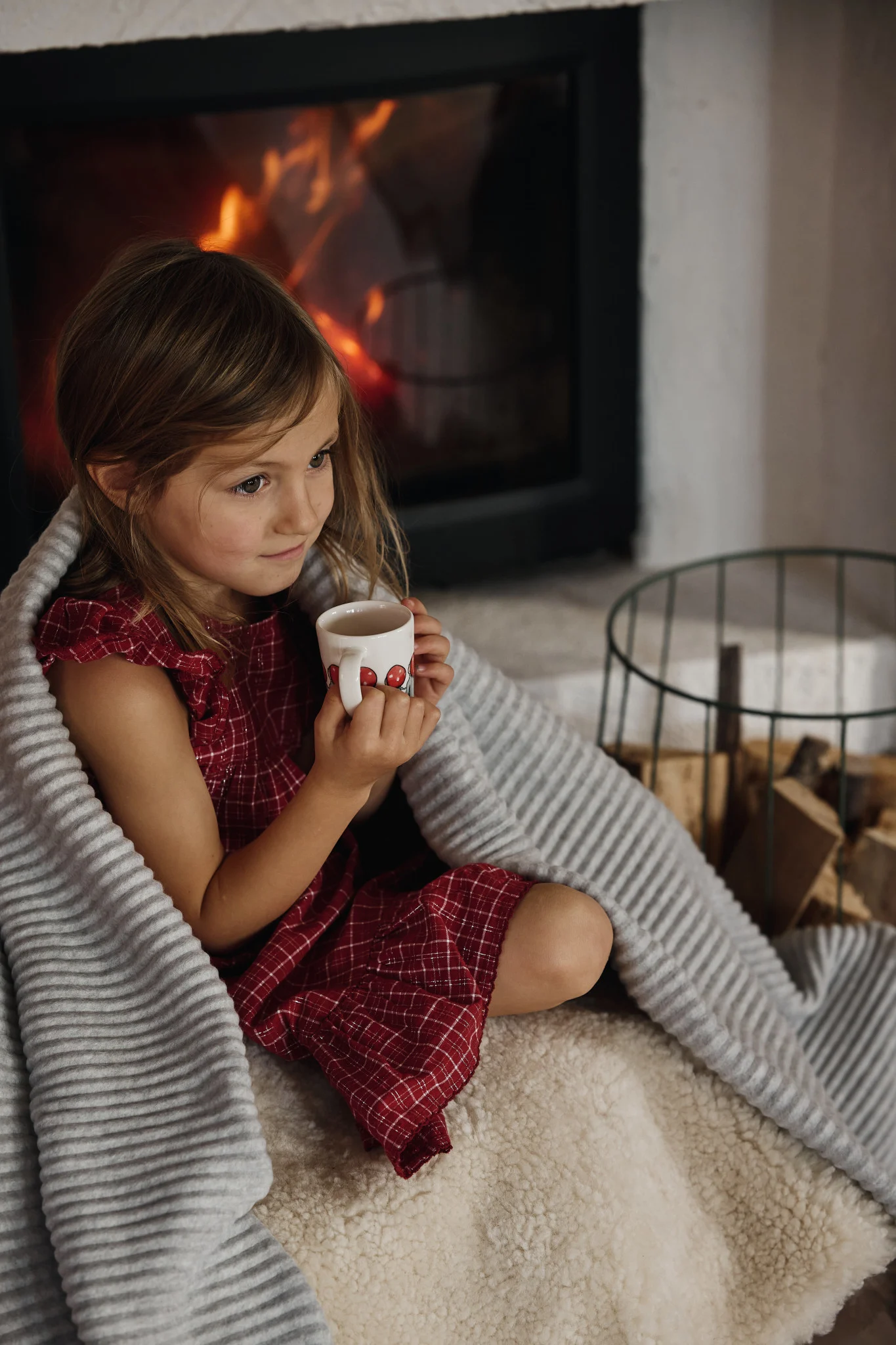 Young girl in a red dress wrapped in a grey blanket, holding a mug by a fireplace with a burning fire.