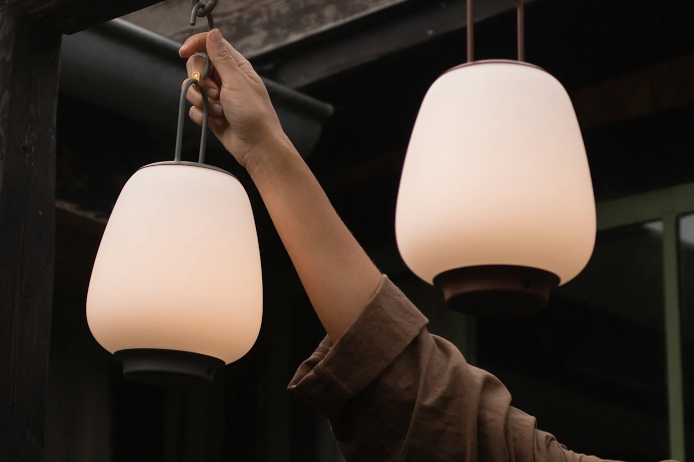 A person's hand holds a glowing white portable lantern with a dark base; another similar lantern hangs nearby.