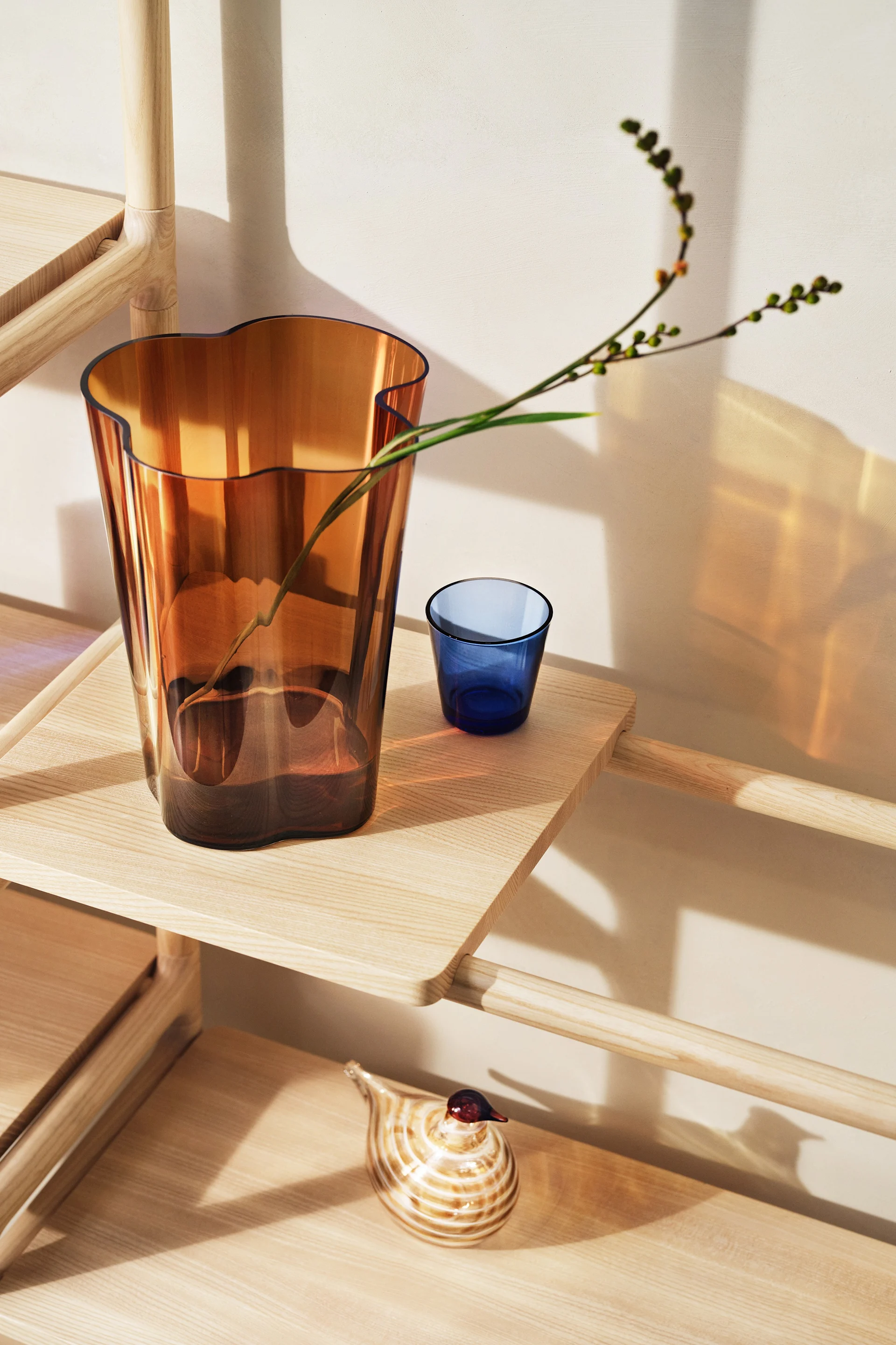 Amber glass vase with wavy top holding a branch, blue glass tumbler, and striped glass bird on a light wood shelving unit.