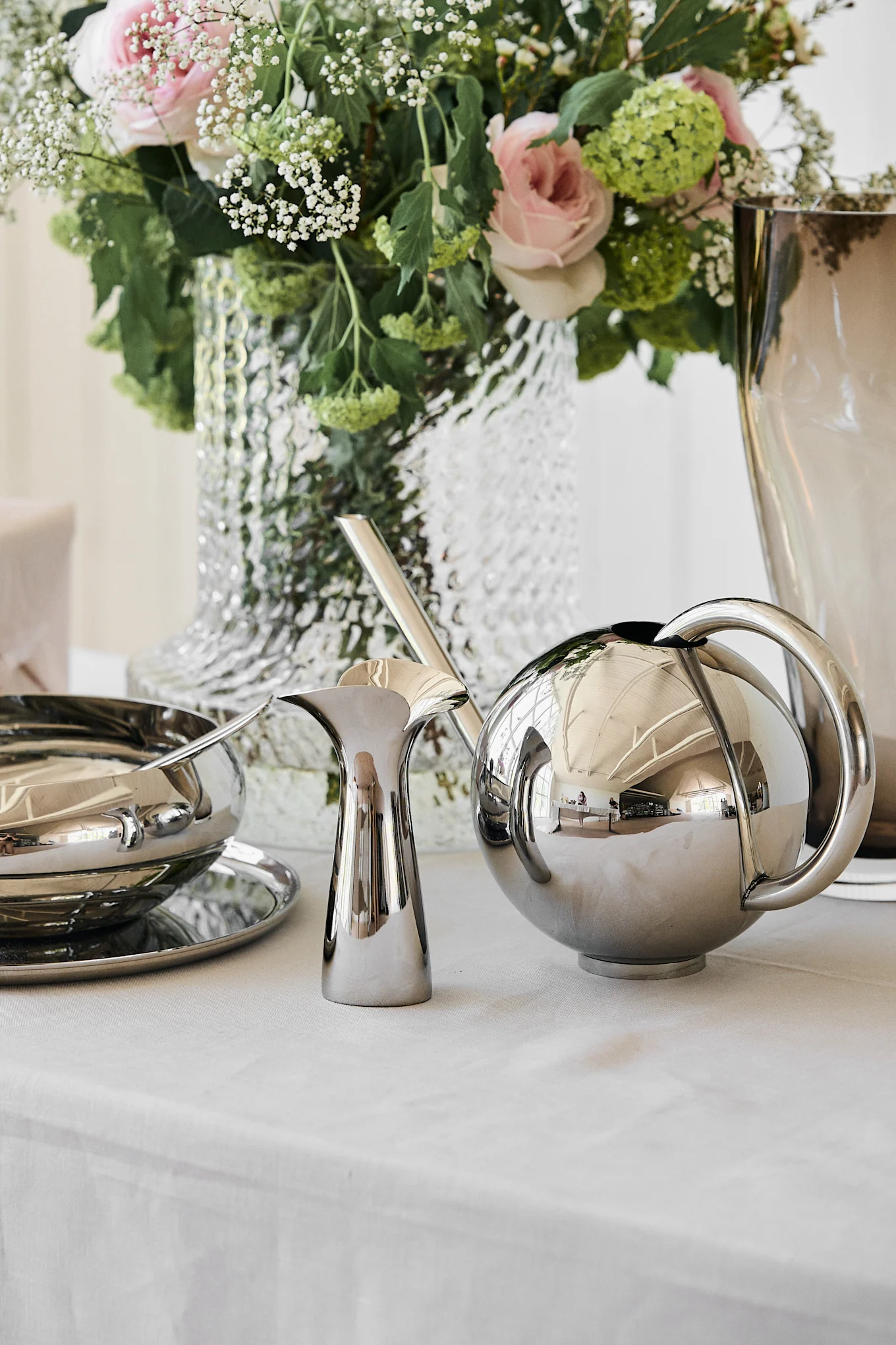 Silver serving set with a bowl, pitcher, and spherical pot on a white tablecloth, next to a floral arrangement with pink roses.
