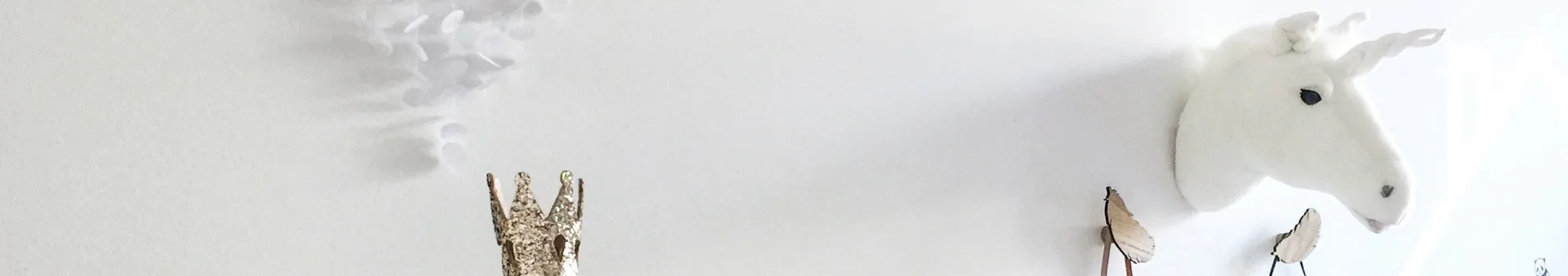 A white plush unicorn head mounted on a white wall, with a gold glitter crown and wooden leaf decorations.
