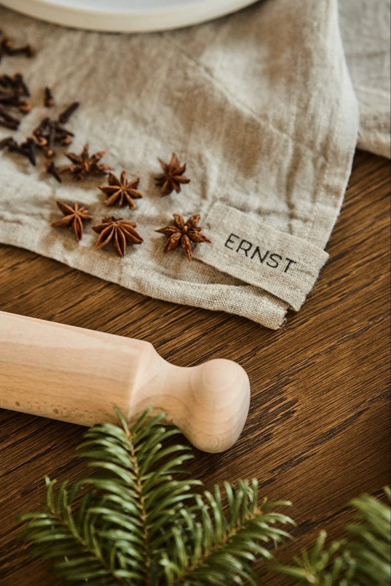 A wooden rolling pin, scattered star anise and cloves, a linen cloth with 'ERNST', and a pine branch on wood.