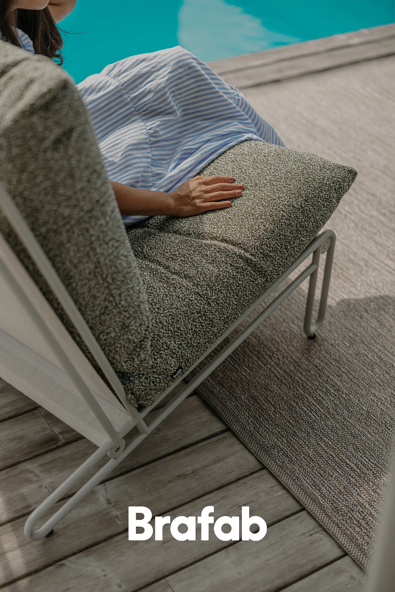 Person sitting on a modern outdoor lounge chair with a dark green speckled cushion on a wooden deck by a pool.