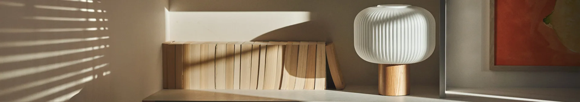 A modern white ribbed lamp with a wooden base stands next to a row of light wooden blocks, with sunlight casting horizontal shadows.
