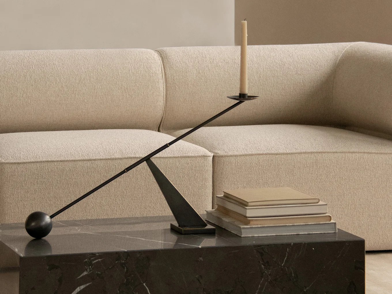 A modern black candelabra with a candle on a dark marble table, beside stacked books and a beige sofa.