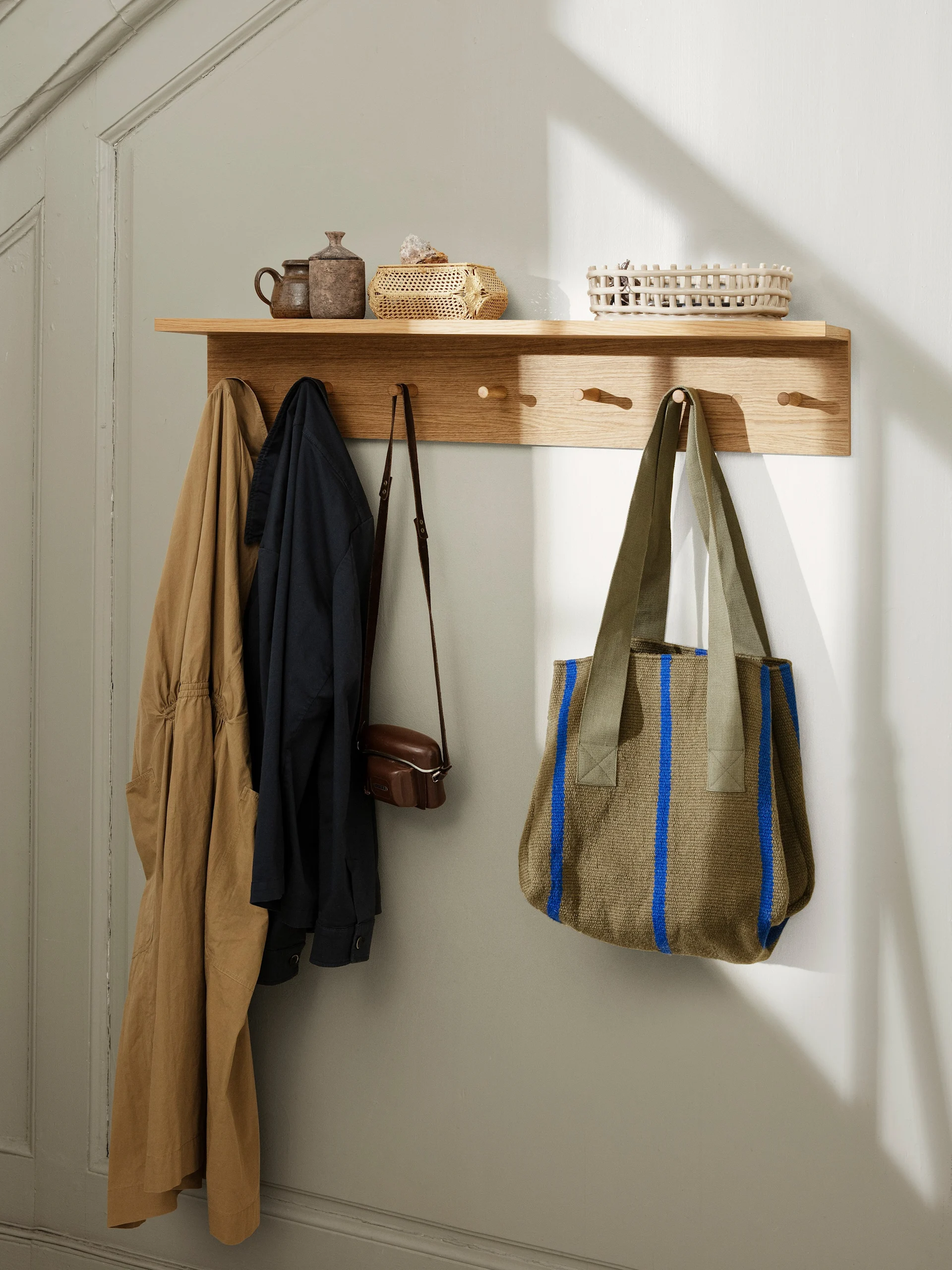 Wooden wall shelf with decorative pots, baskets, and coats, camera bag, and olive green striped tote bag hanging from pegs.