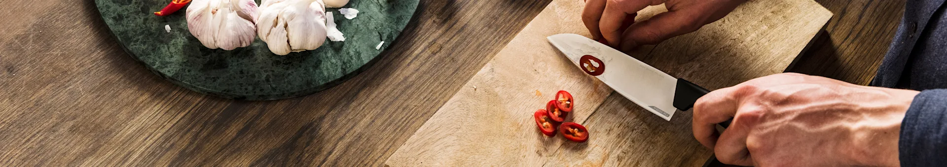 Hands slicing red chili peppers on a wooden board, with garlic and a chili on a marble surface.