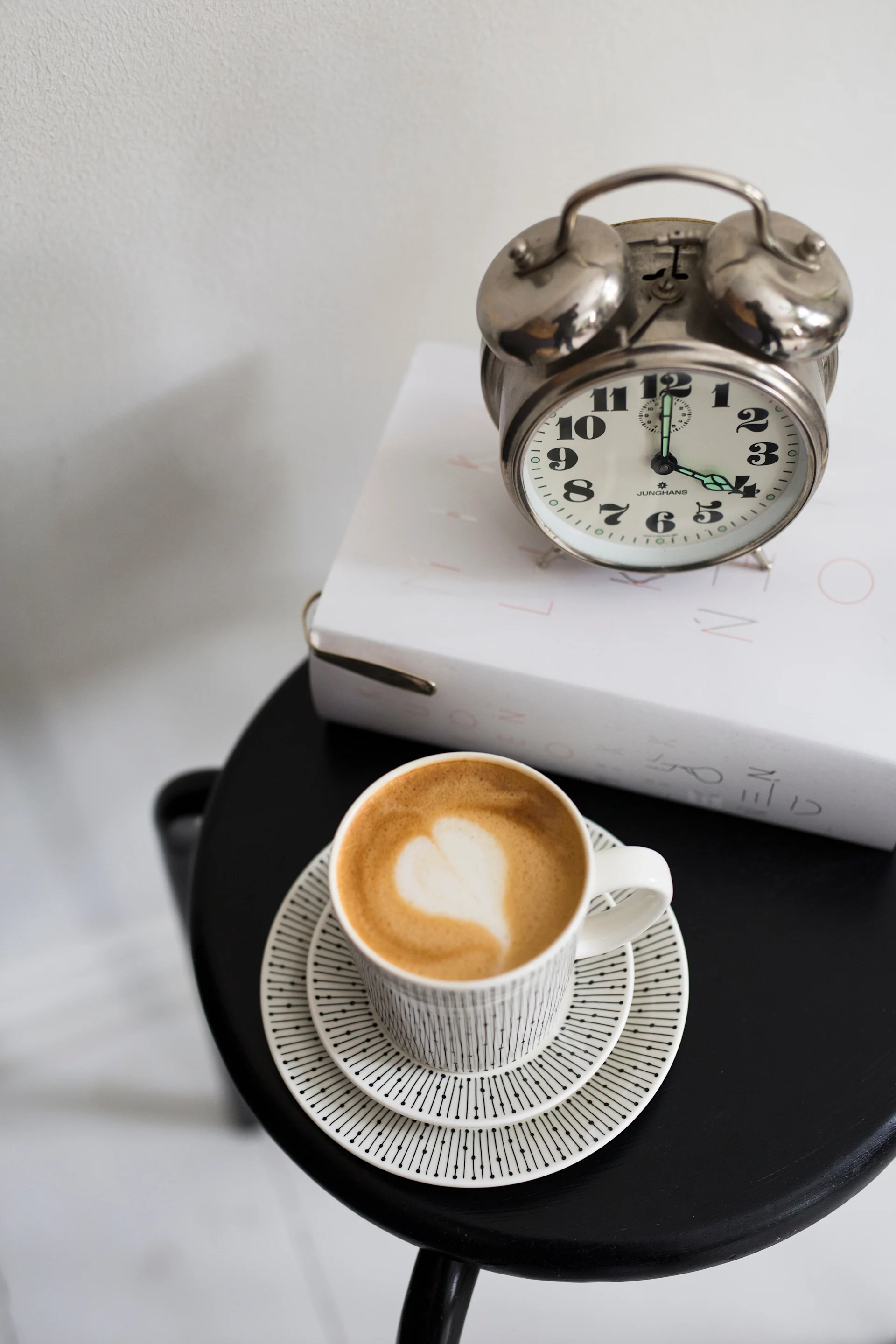 A latte with heart art in a striped cup on saucers, next to a silver alarm clock on white books.