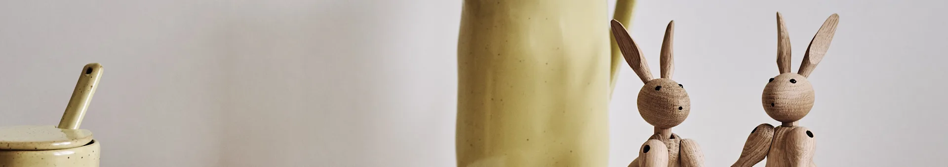 A light yellow ceramic jar with a spoon, a pitcher, and two wooden bunny figurines.