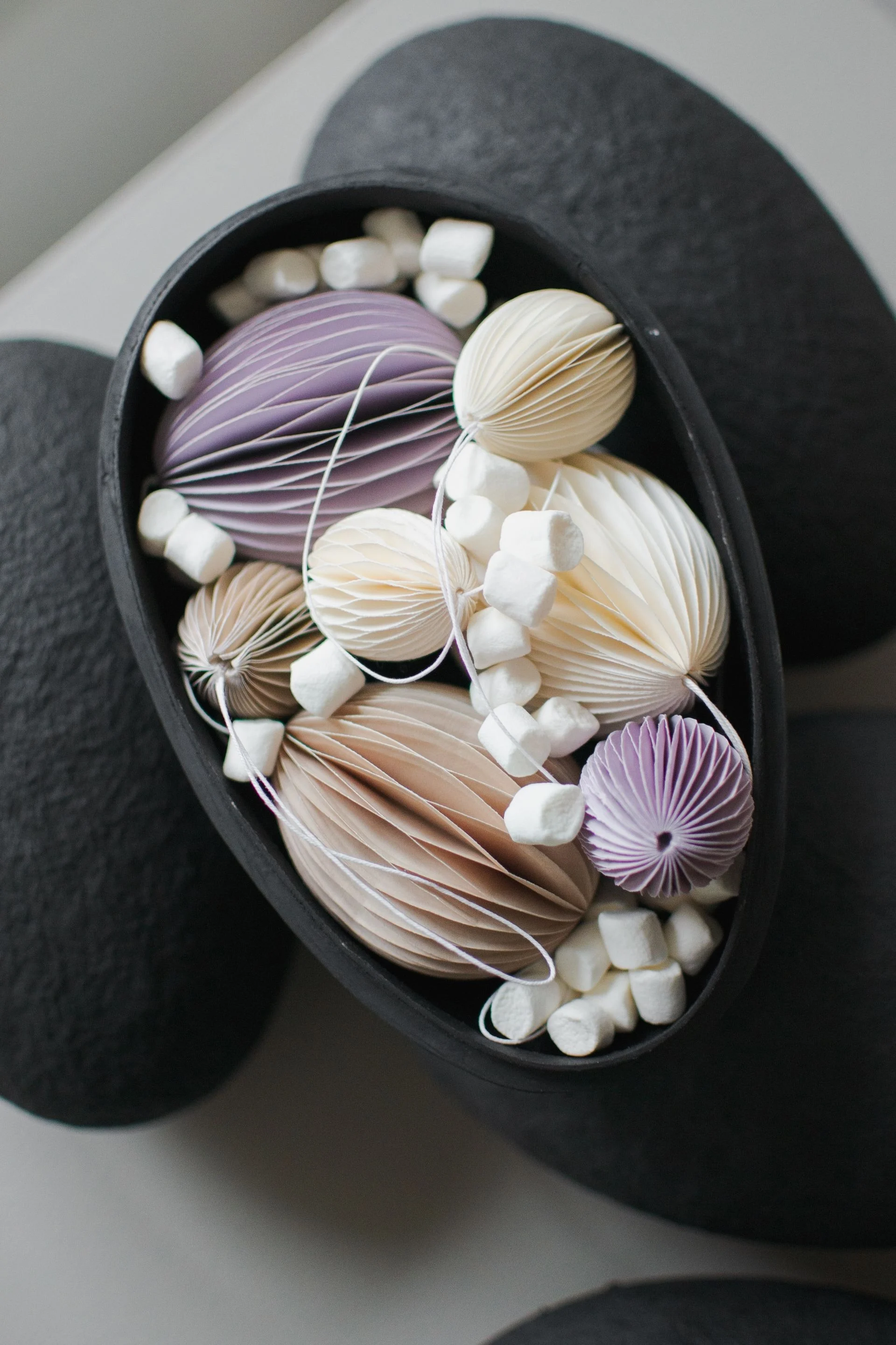 Elongated black bowl filled with accordion-folded paper decorations in pastel lavender, cream, and beige, alongside white mini marshmallows.