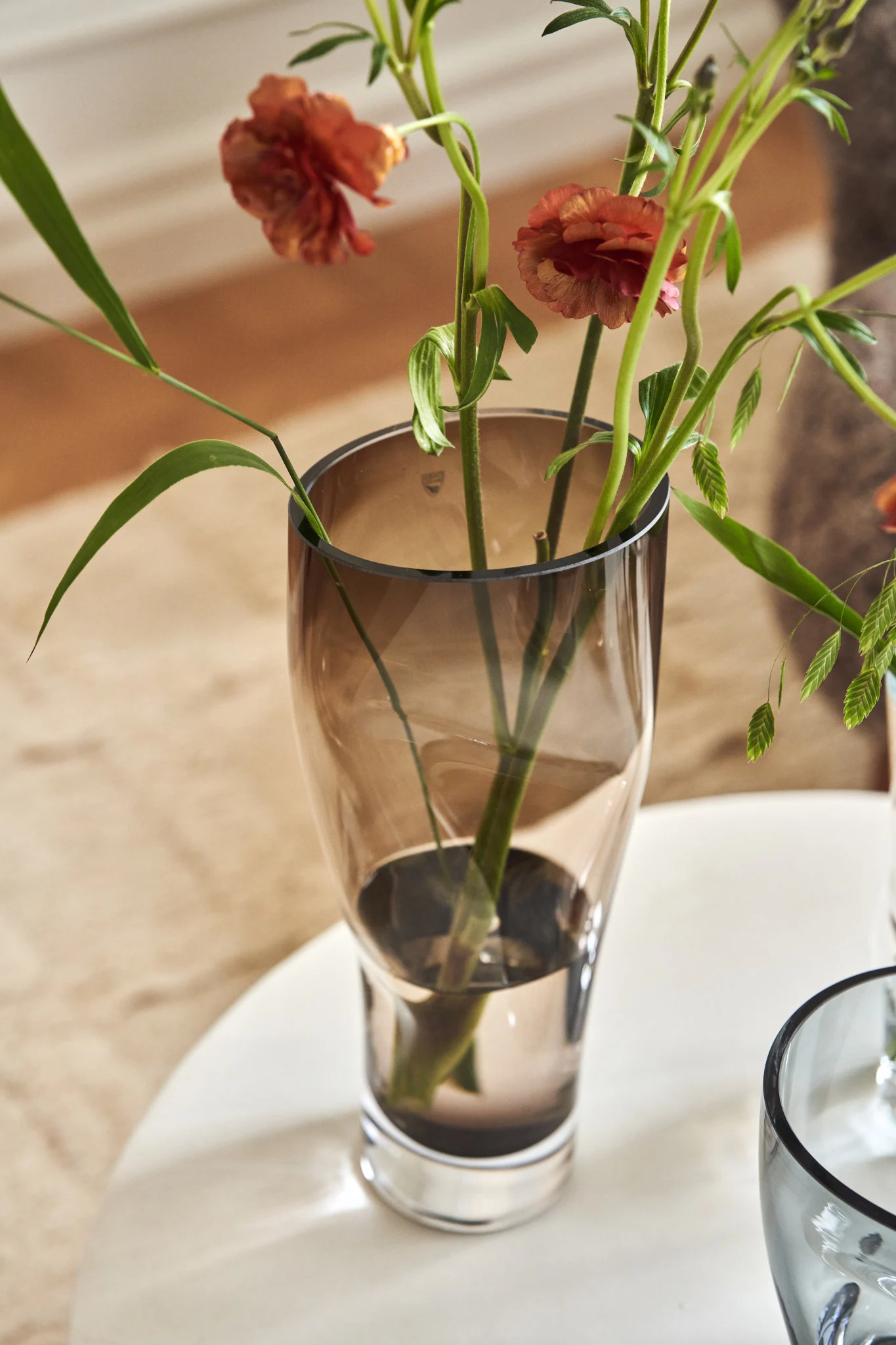 Smoky brown glass vase with orange ranunculus flowers and green foliage on a white table.