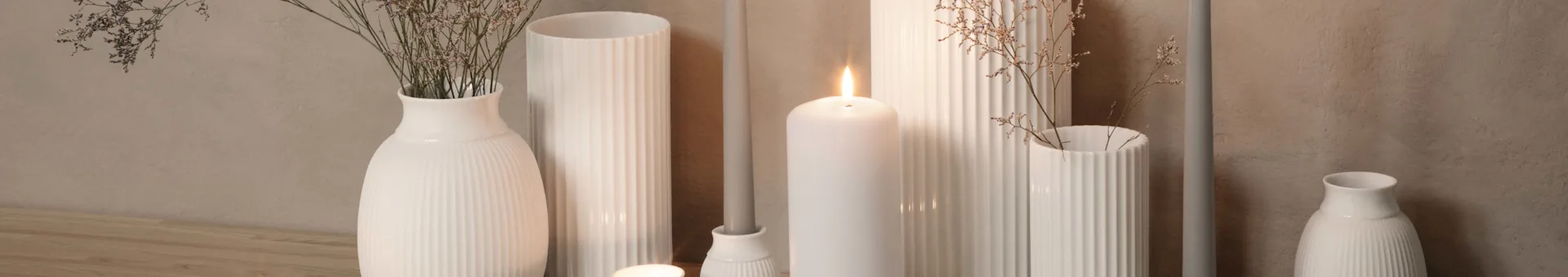 Arrangement of white ribbed vases and candle holders with dried flowers and a lit white pillar candle on a wooden surface.