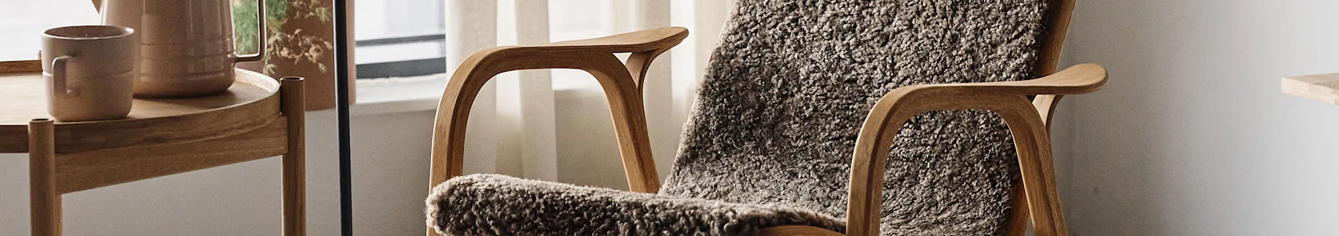 Wooden armchair with a fuzzy gray throw, next to a wooden side table holding a beige mug and carafe.