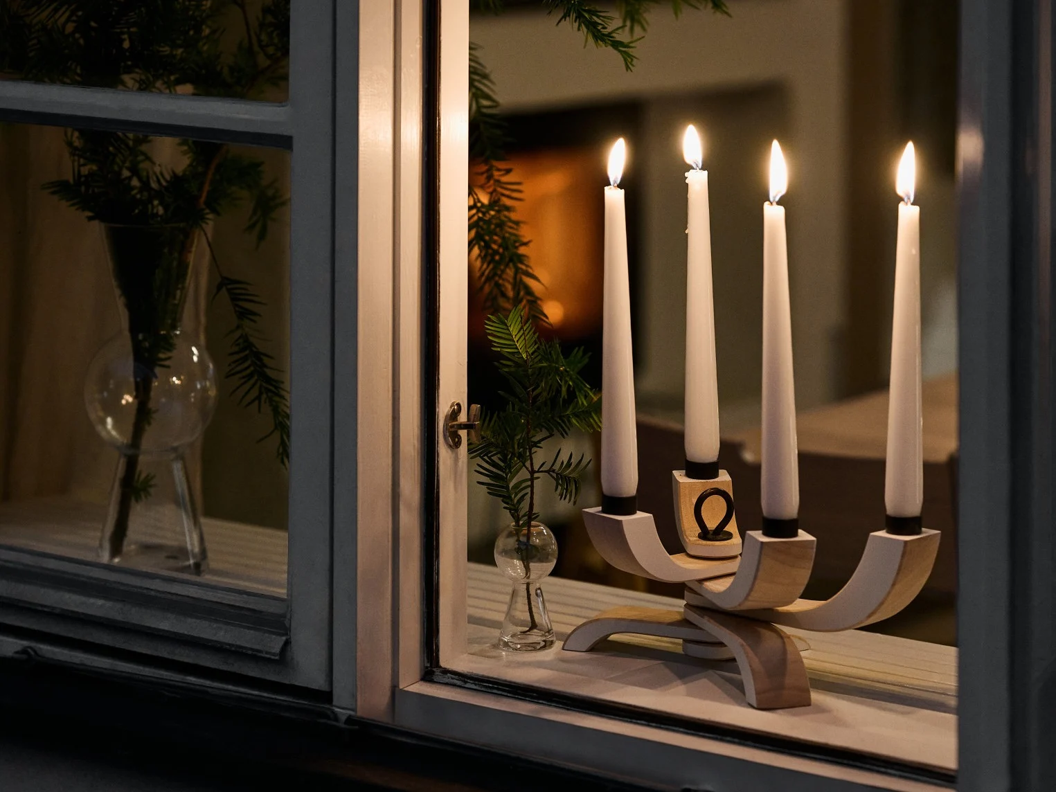 A modern wooden candle holder with four lit white candles sits on a window sill next to small vases with green branches.