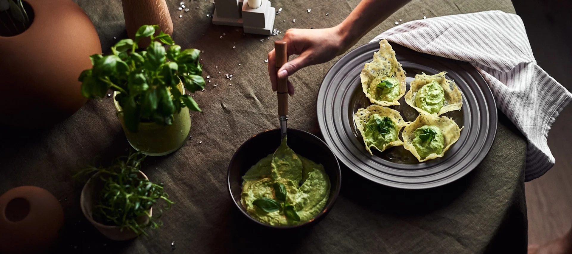 Hand serving green dip into crispy cheese cups, alongside fresh basil and herbs on a dark green table.