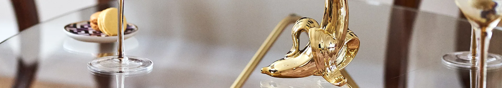 A glossy golden banana-shaped decorative object on a glass table with macarons and gold-stemmed glasses.