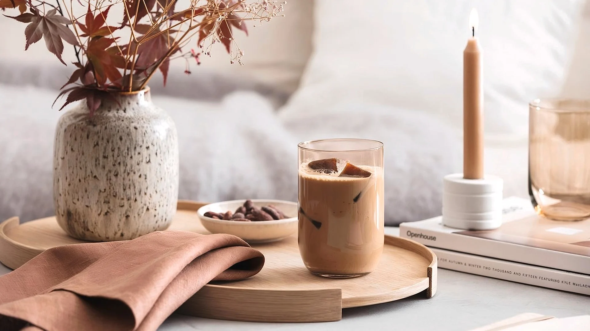 A wooden tray with an iced coffee, a speckled vase of dried leaves, a bowl of nuts, and a terracotta napkin, with a lit candle.