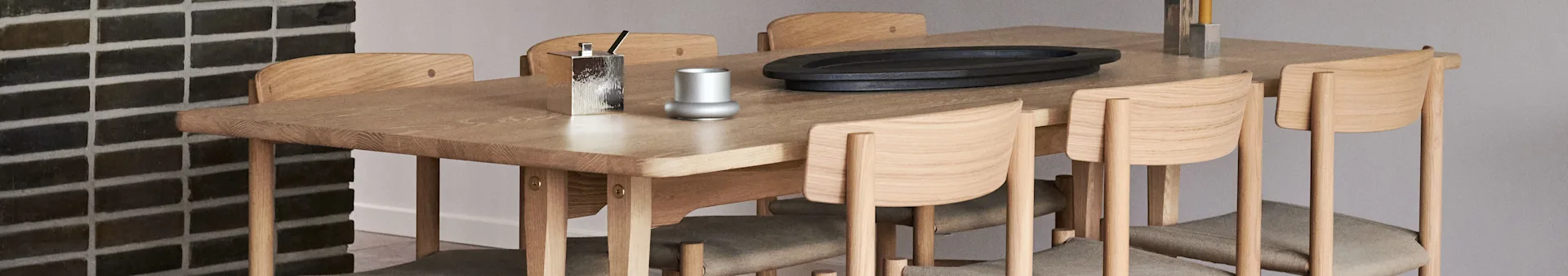 Light wooden dining table with upholstered chairs. On top: silver container, small vase, black oval tray, two silver candle holders.