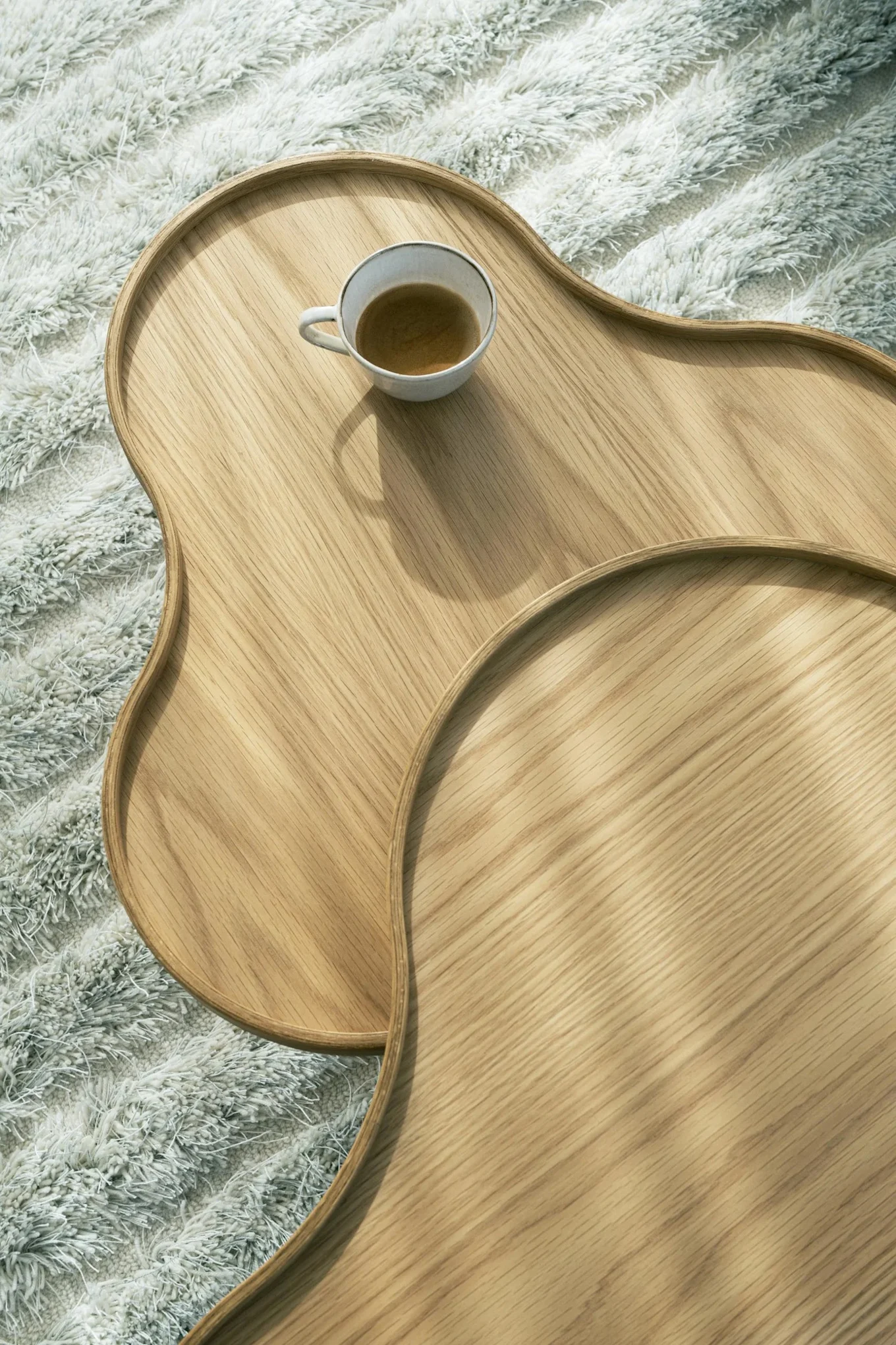 Overhead view of two wavy wooden trays, one with a coffee cup, on a light shaggy rug.