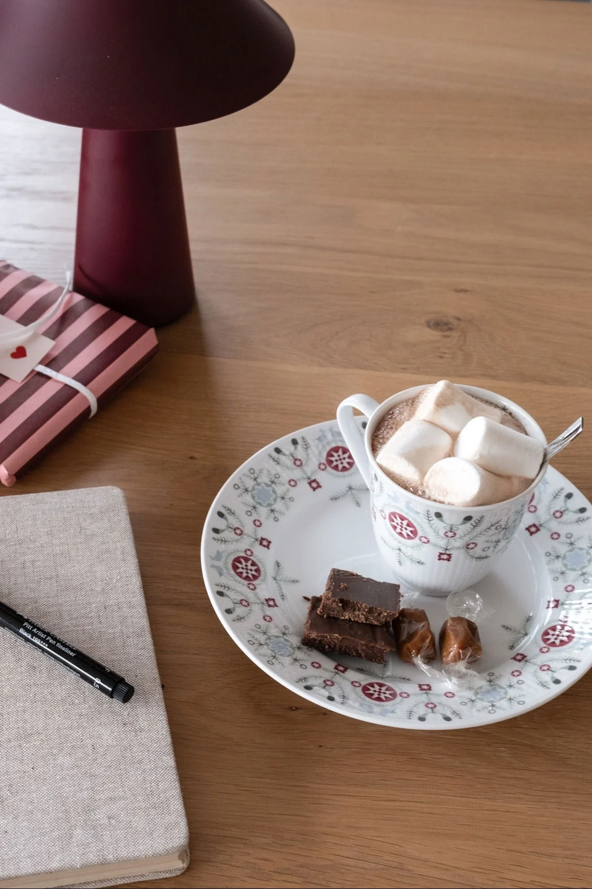 Hot chocolate with marshmallows, chocolate fudge, and caramels on a festive plate with a gift, notebook, and lamp.