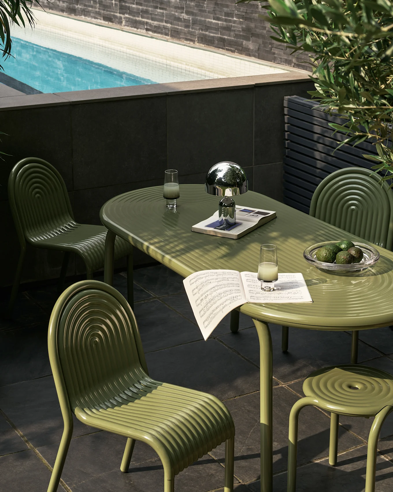 Outdoor patio with olive green ribbed dining set by a pool. Table has silver lamp, drinks, music, and avocados.