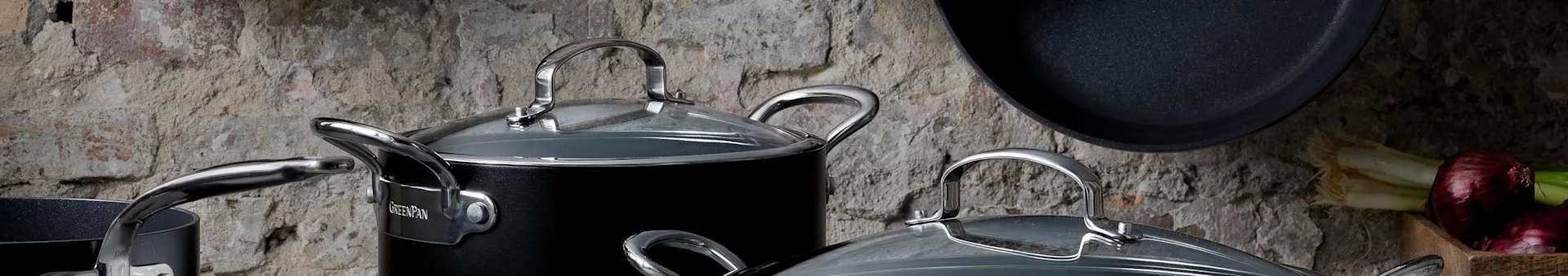 Assortment of black GreenPan pots and pans with silver handles and glass lids in a rustic kitchen setting.