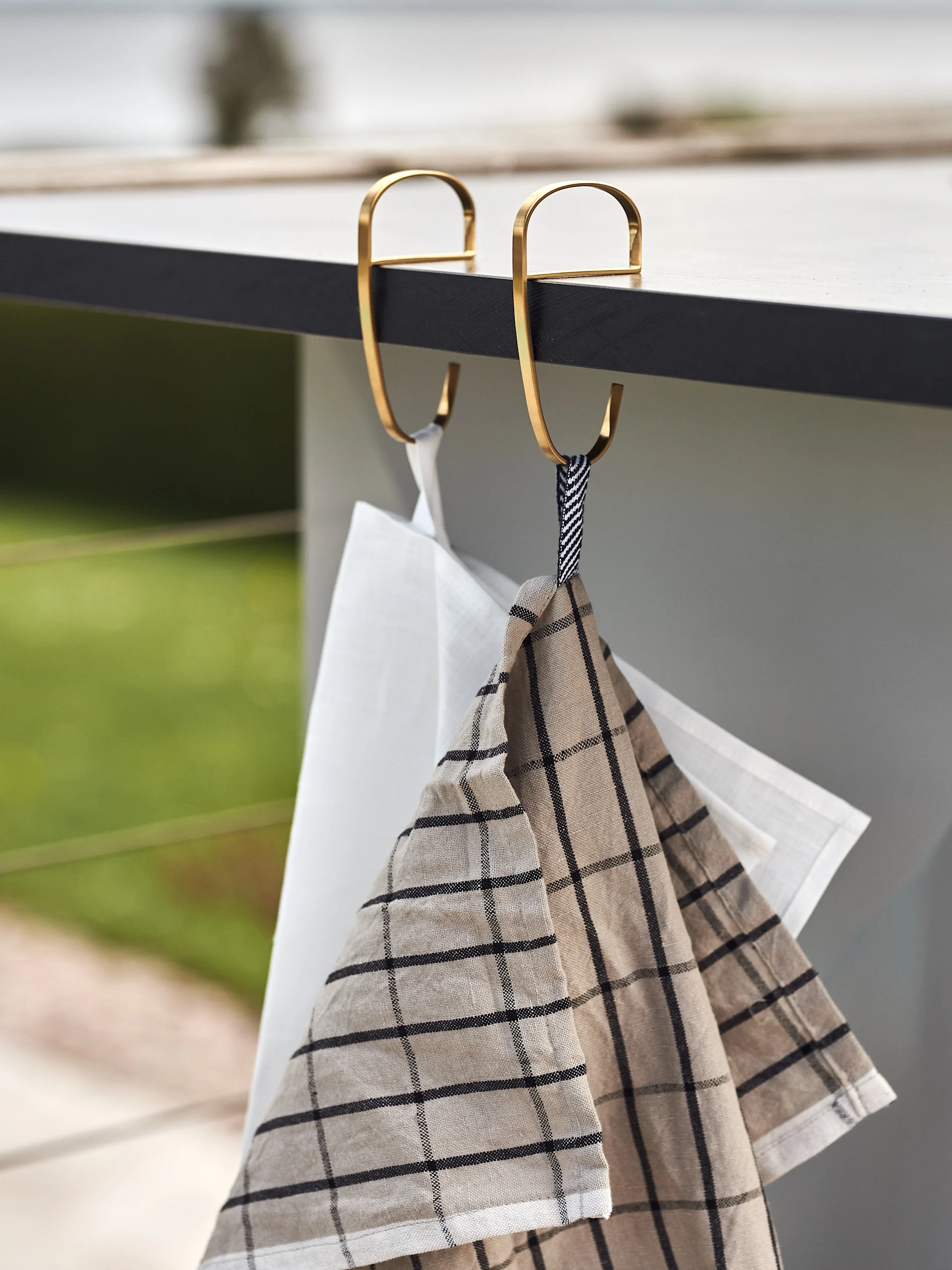 Two kitchen towels, one white and one beige plaid, hang from golden S-hooks on a dark counter.