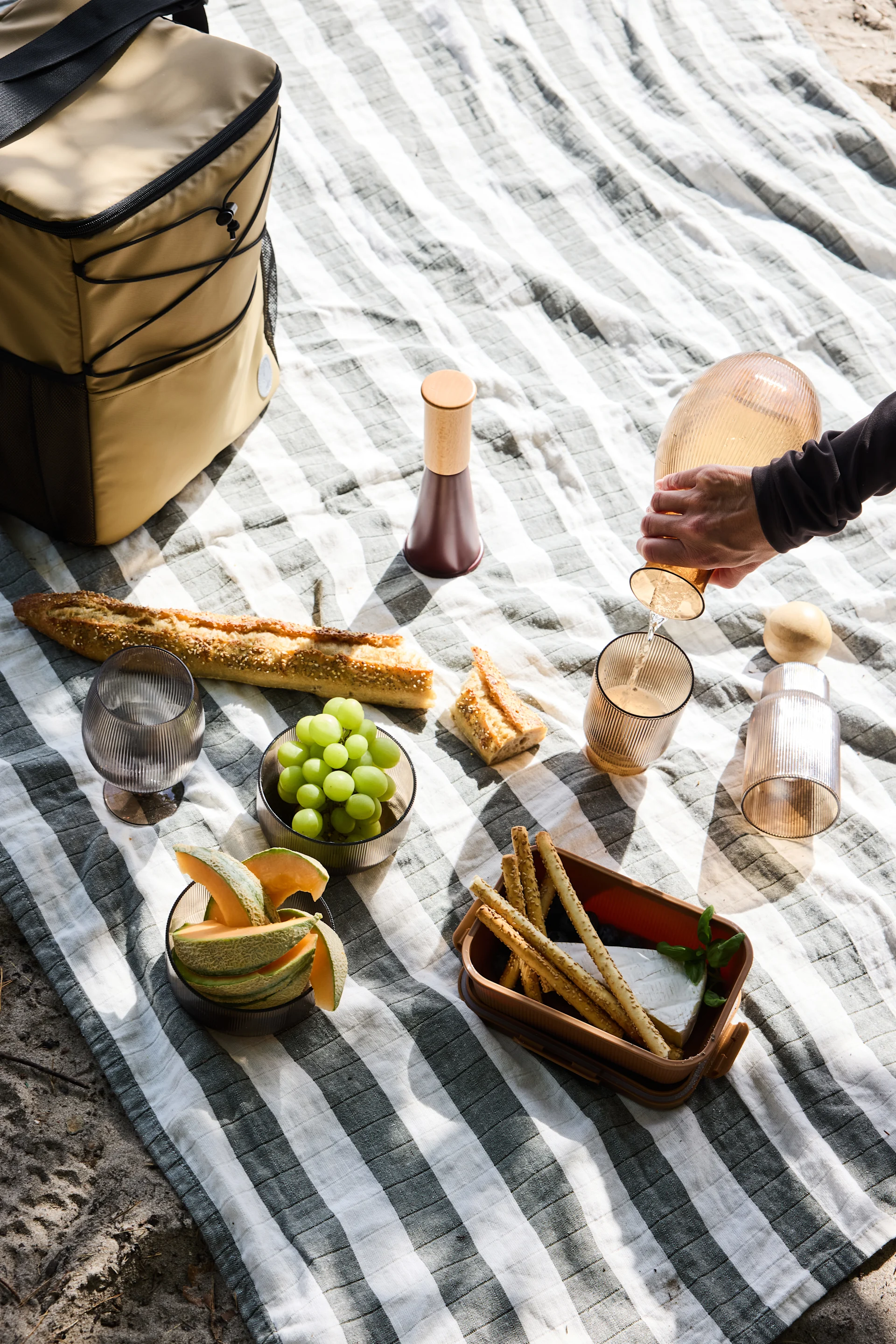 Overhead view of a picnic spread on a striped blanket, with a person pouring from a fluted carafe, baguette, fruit, cheese, and breadsticks.