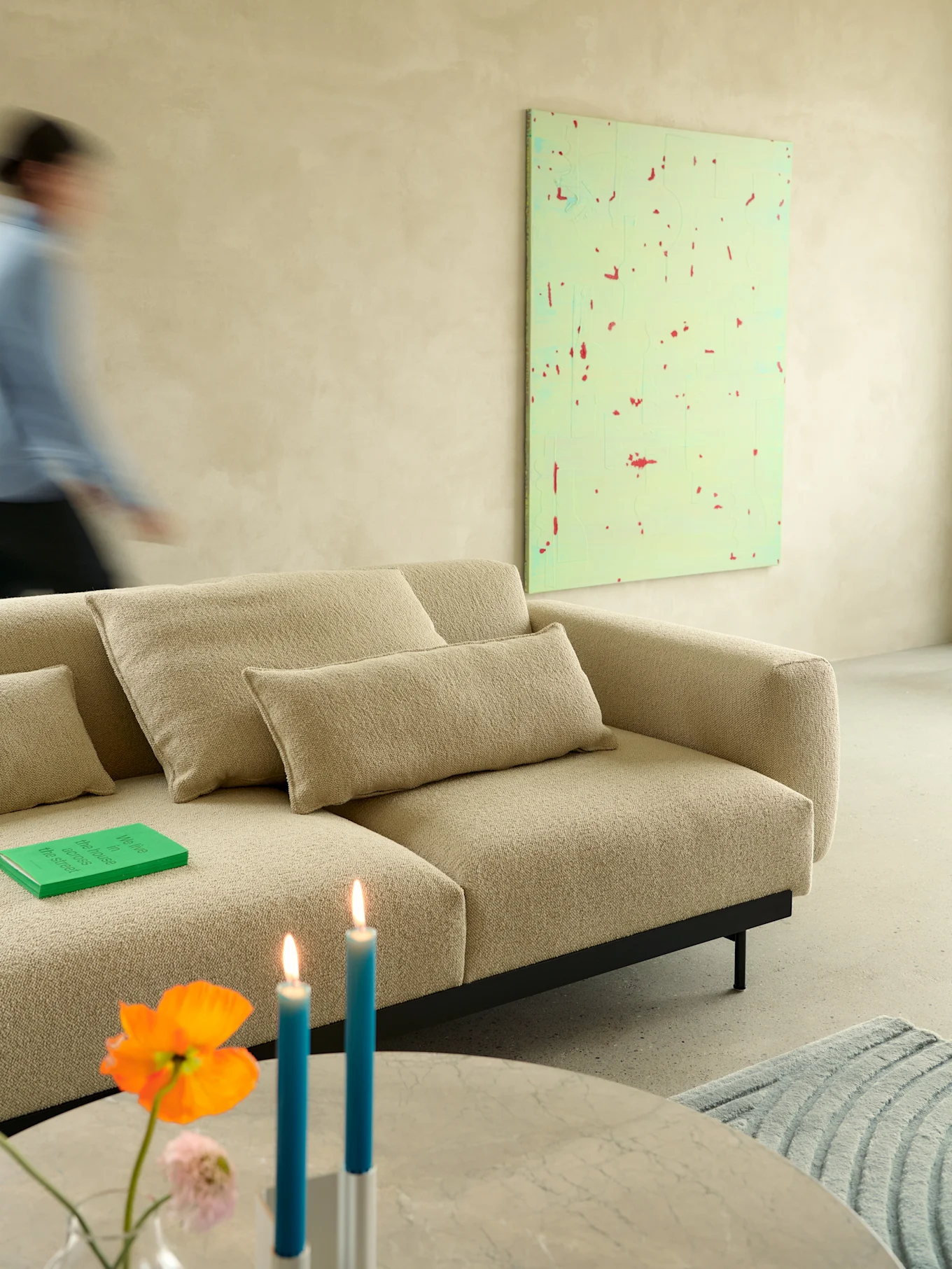 Beige textured sofa in modern room. Blurred person walks past. Green book, lit blue candles, and flowers on marble table.
