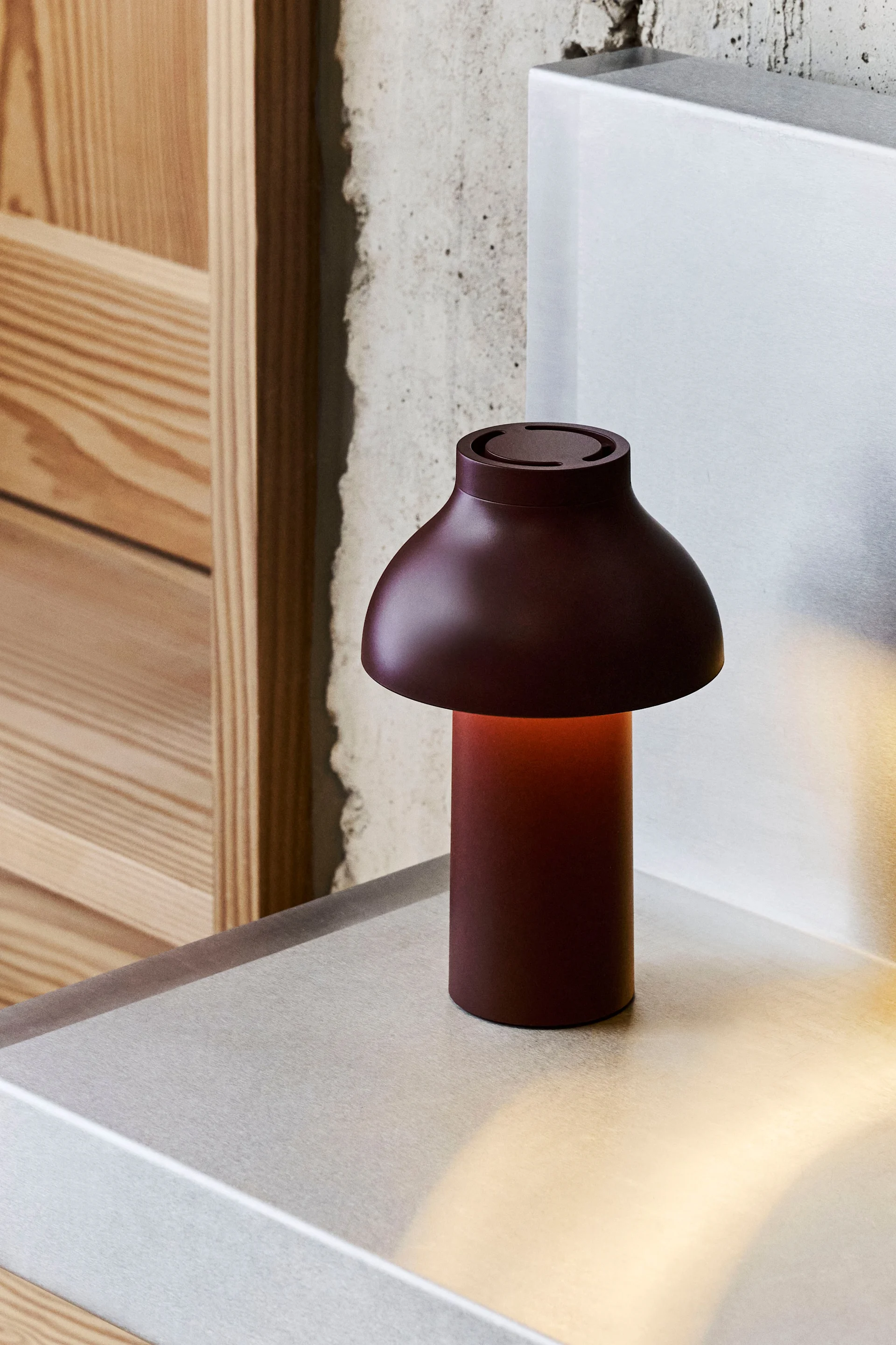 A dark red mushroom-shaped table lamp with a warm glow sits on a metallic surface, with concrete and wood in the background.