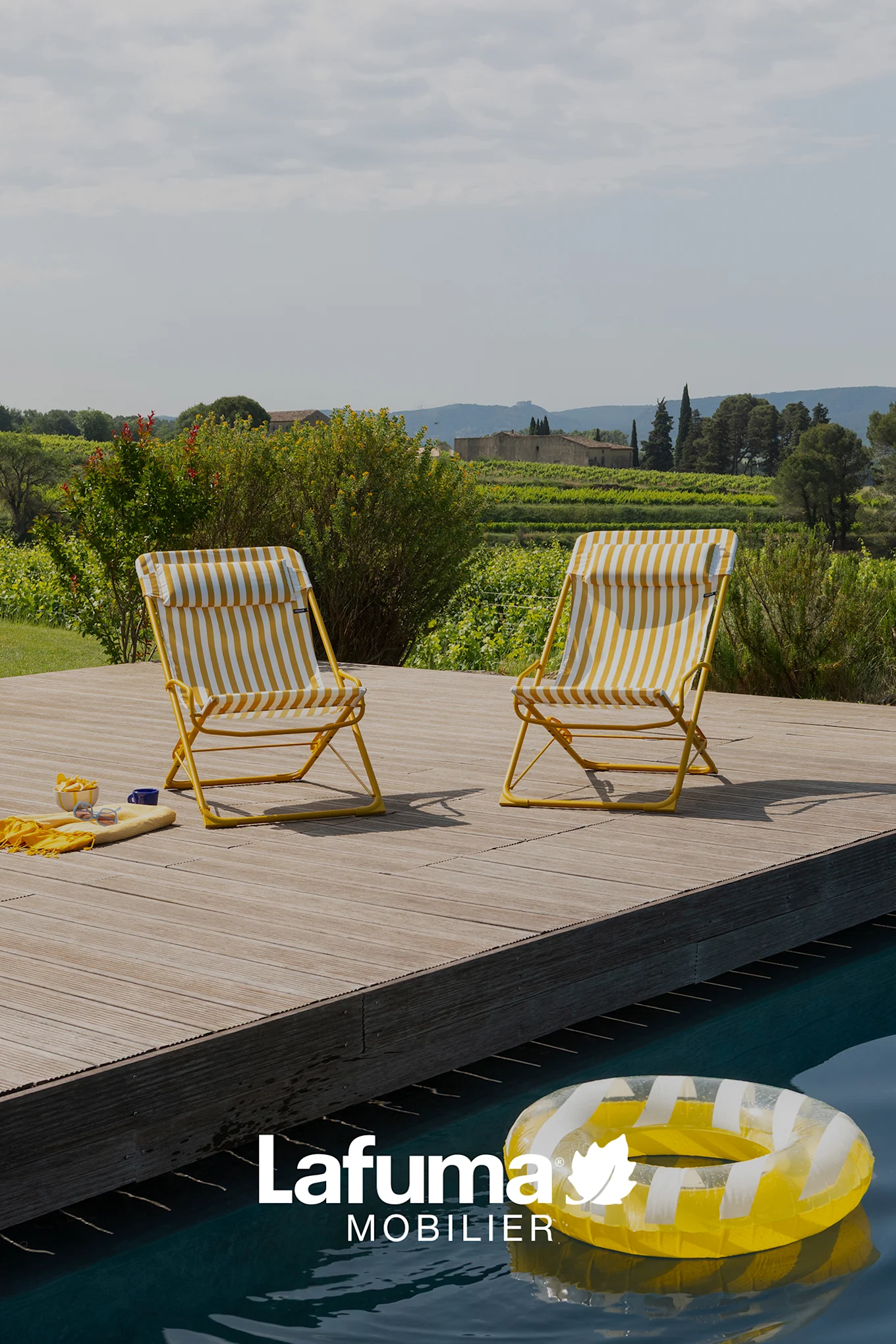 Two yellow and white striped folding chairs by a pool, with a vineyard and buildings in the background.