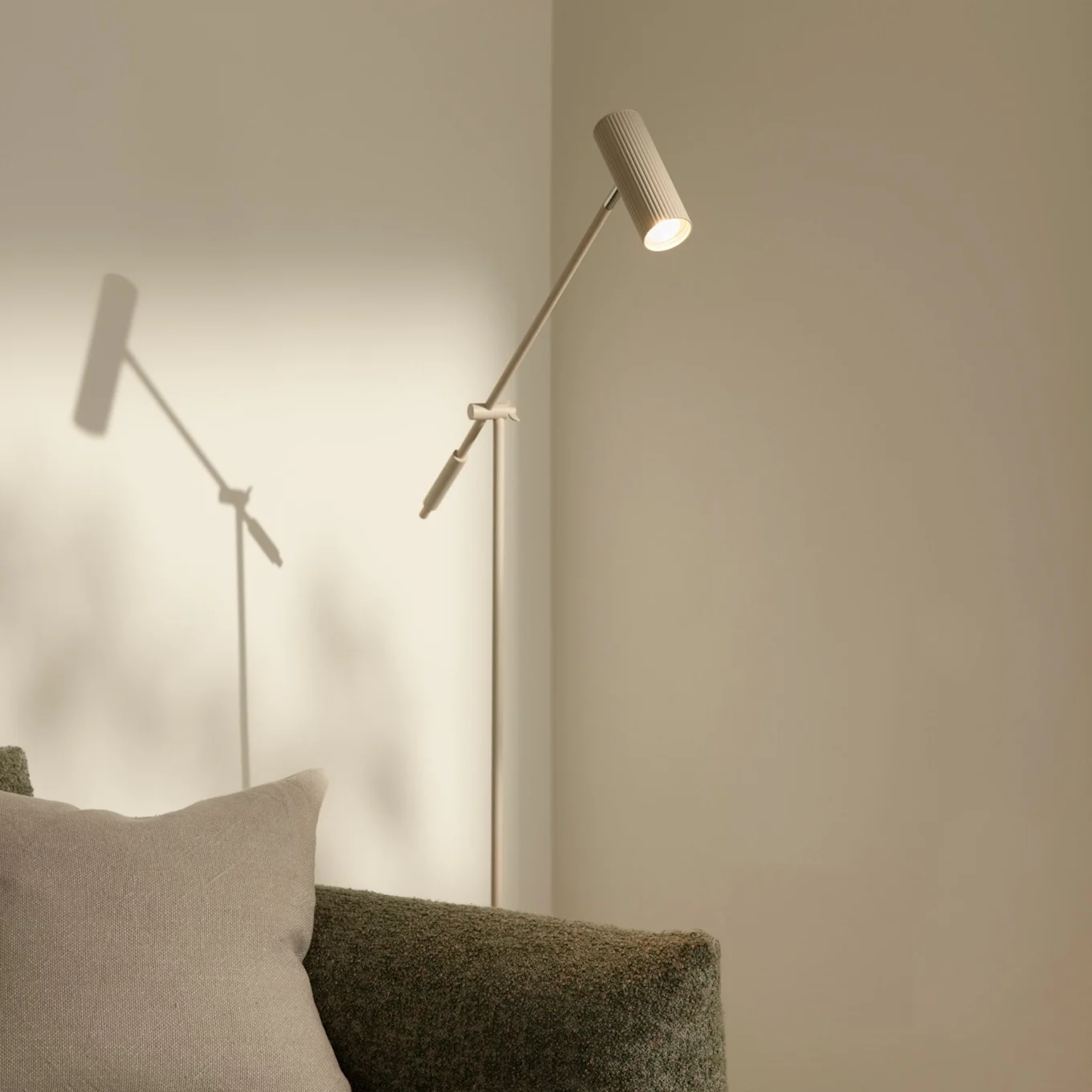 A modern, slender floor lamp with a ribbed head illuminating a light wall, next to a green textured sofa with a beige pillow.