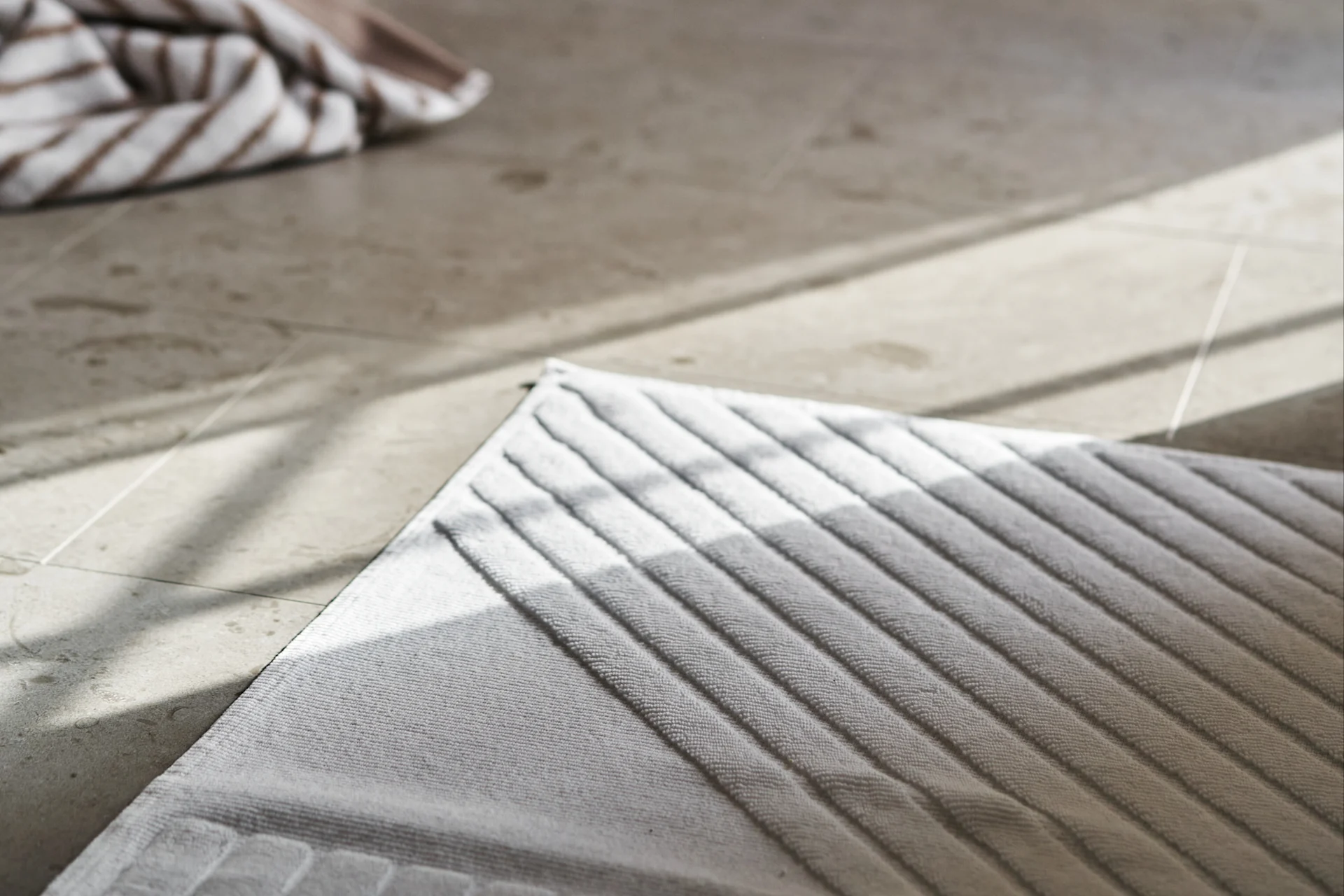 Light gray ribbed mat on tiled floor with diagonal sunlight and striped towel in background.