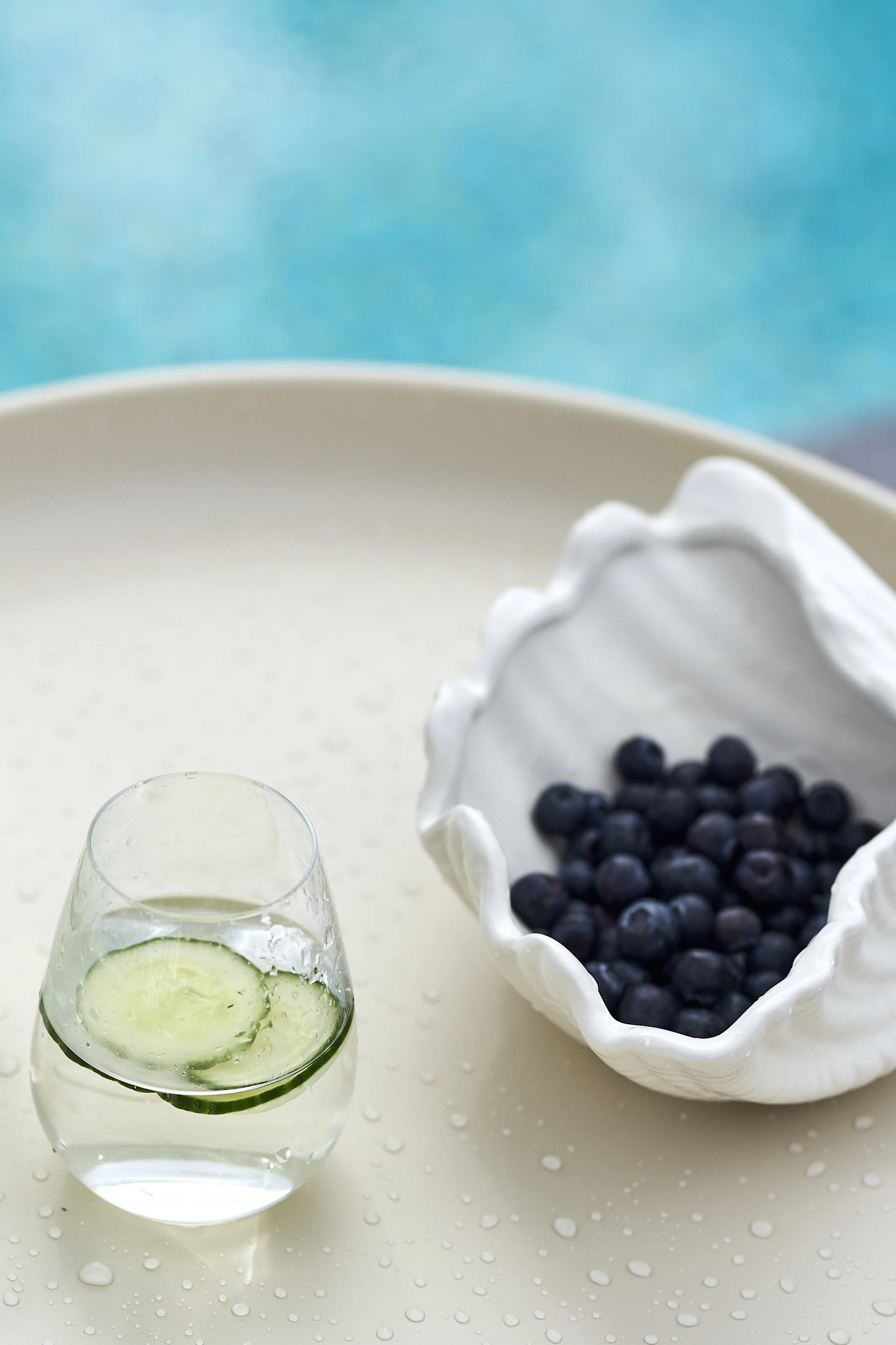 Cucumber water and blueberries in a shell-shaped bowl on a wet table next to a pool.