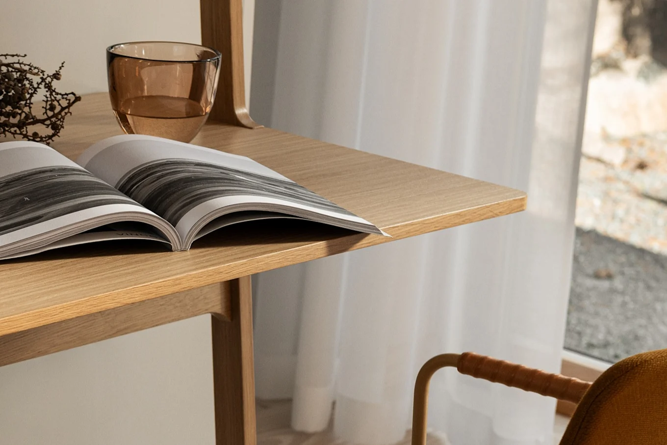 An open magazine and a glass of water on a light wooden desk, with a chair and sheer curtain in the background.