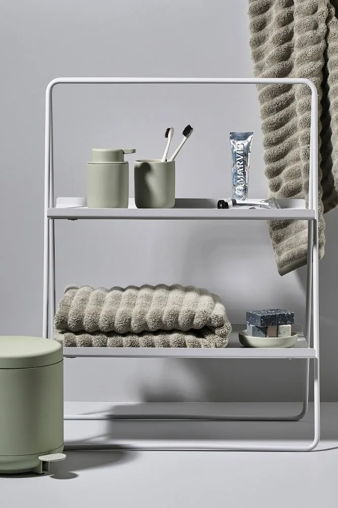 White 3-tier bathroom shelf with sage green accessories: soap dispenser, toothbrushes, toothpaste, folded textured towels, and soap bars.