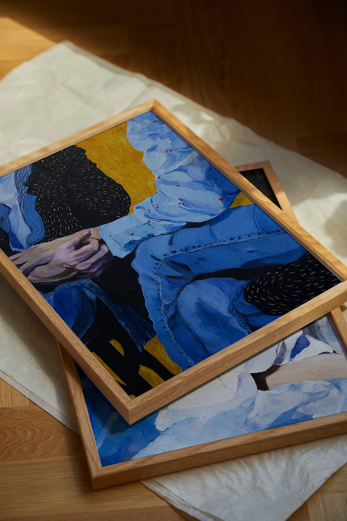 Two framed oil paintings, the top one showing two people in blue holding hands, on paper on a wooden floor.