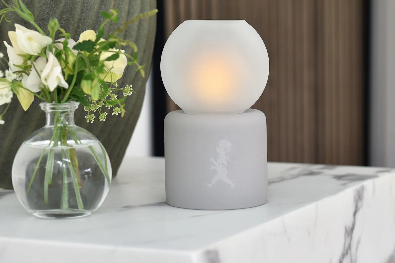 Frosted white spherical lamp with warm glow on cylindrical base featuring walking figure, next to a vase of white and yellow flowers on a marble surface.