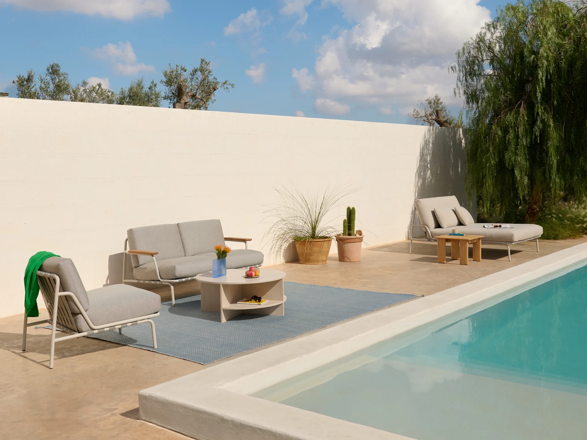 Outdoor patio with light grey sofa, armchairs, coffee table on a blue rug, and a lounge chair by a pool.