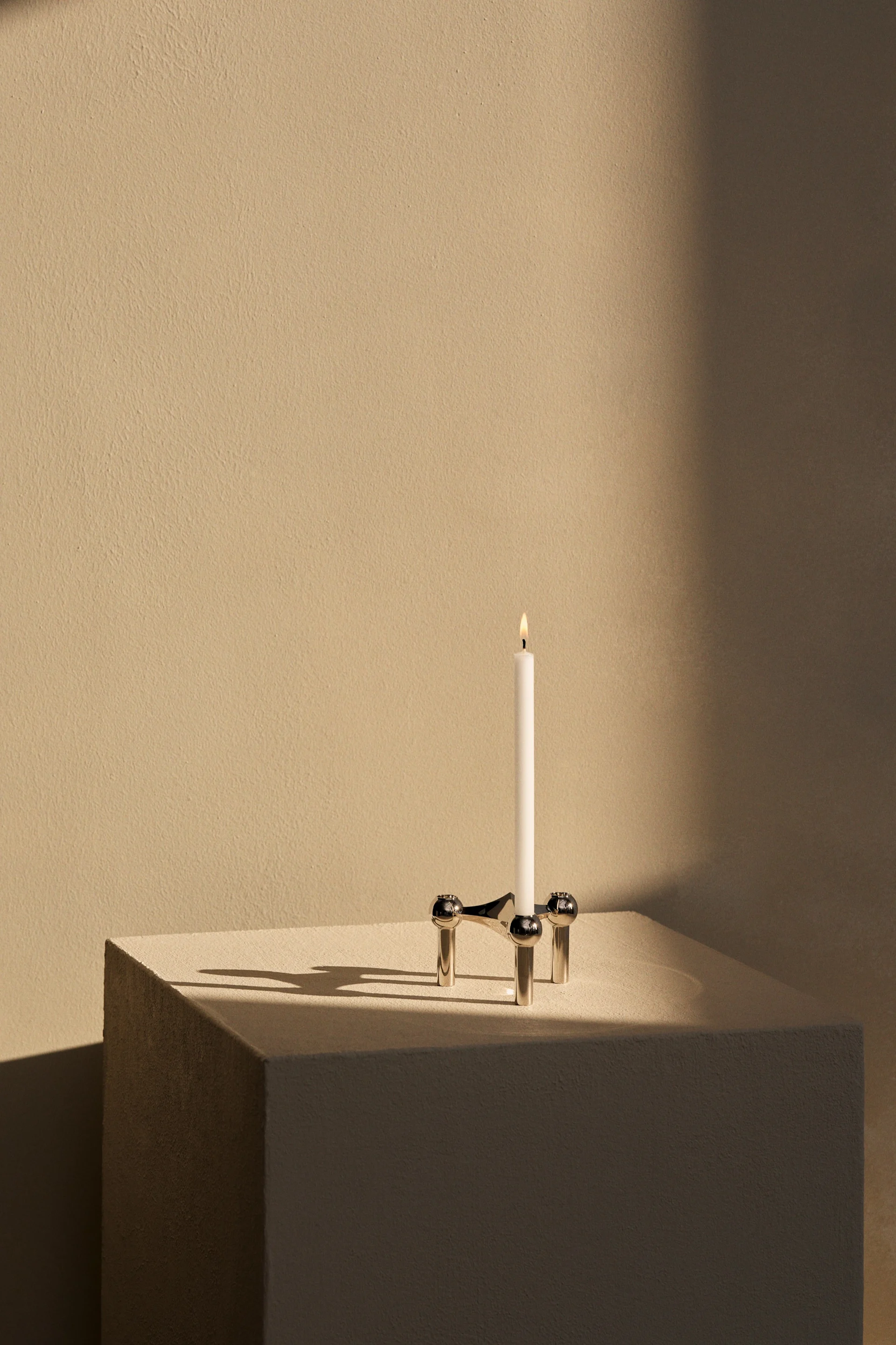 A single white candle burning in a silver, three-legged candlestick with spherical details, casting long shadows on a textured light beige surface.