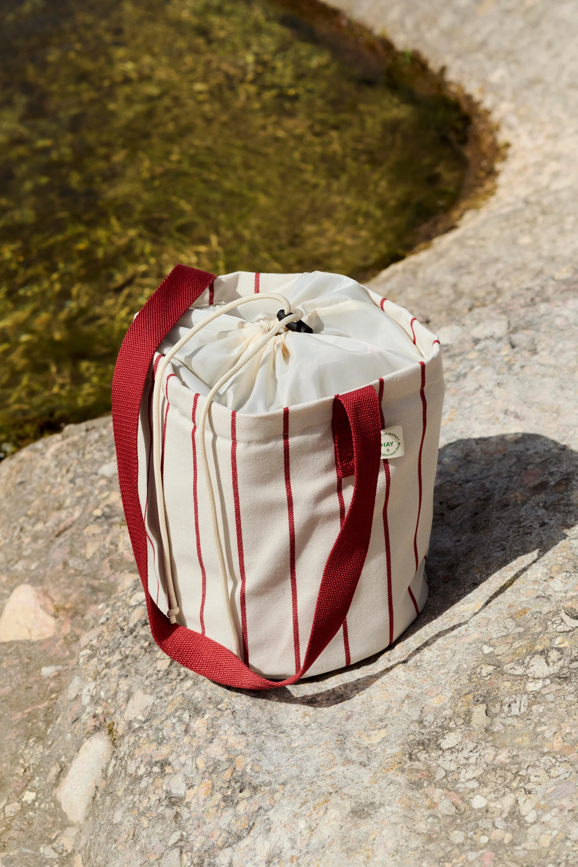 A cylindrical cream bag with red vertical stripes and a drawstring closure, on rocks by water.