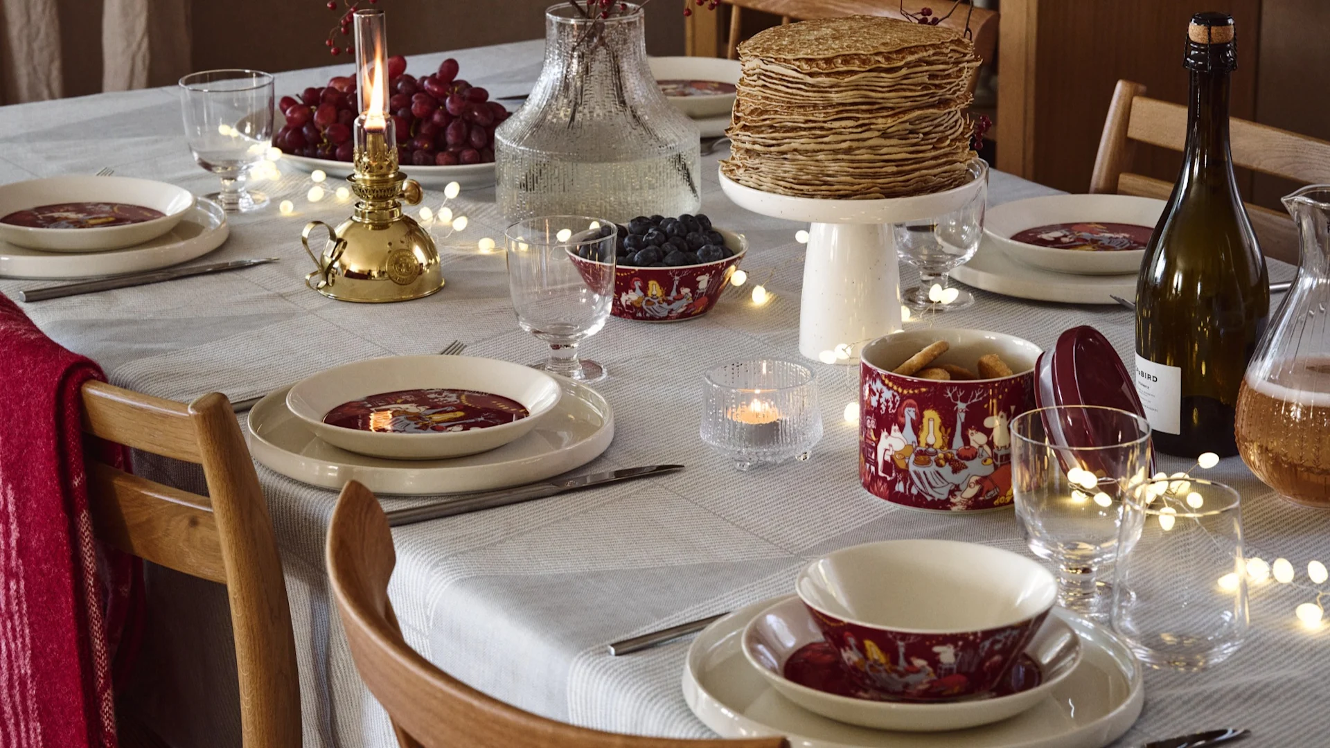 A dining table set with white plates, red patterned bowls, a stack of pancakes, fruit, beverages, and lit decorative lights.
