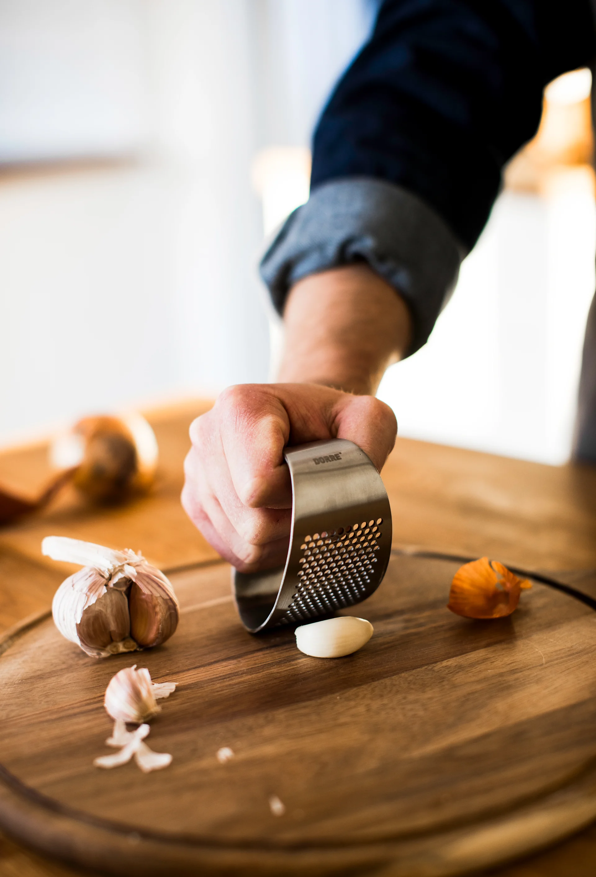 A hand uses a stainless steel garlic press to crush a garlic clove on a wooden cutting board.