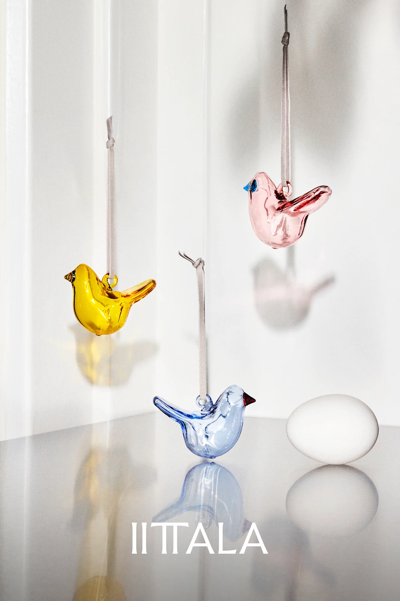 Three colorful glass birds (yellow, pink, blue) hang with a white egg on a reflective surface.