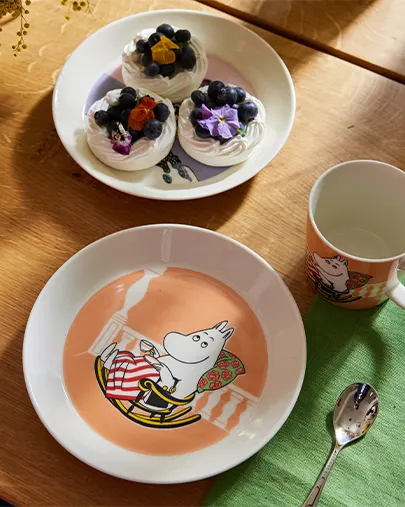 Moomintroll plate, mug, and spoon next to a plate of meringues with blueberries and flowers on a wooden table.