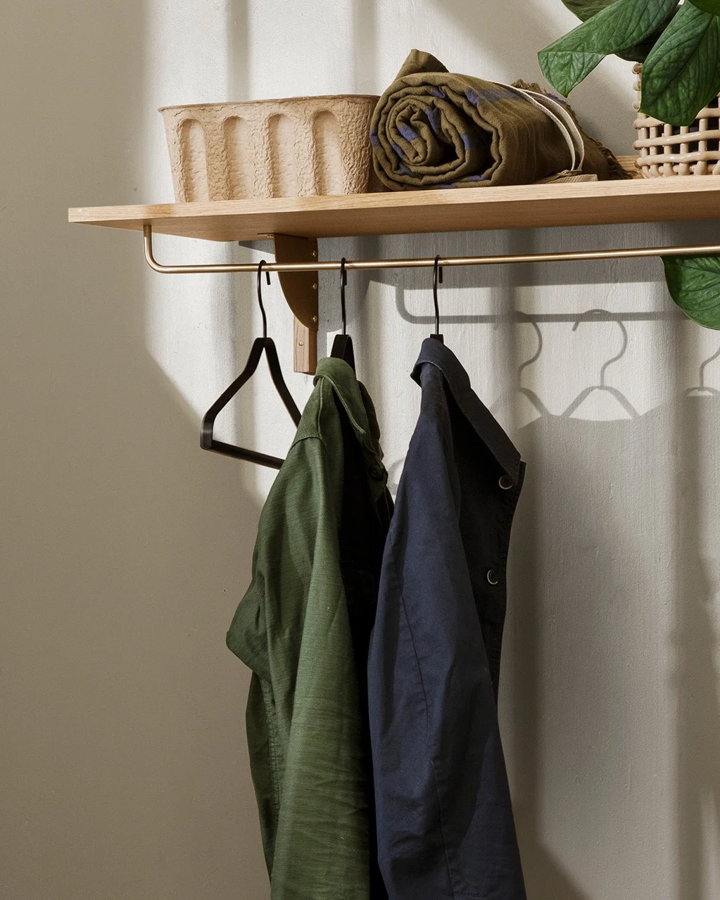 A wooden shelf with a brass rail holding a green and a navy coat, above a textured basket and rolled blanket.