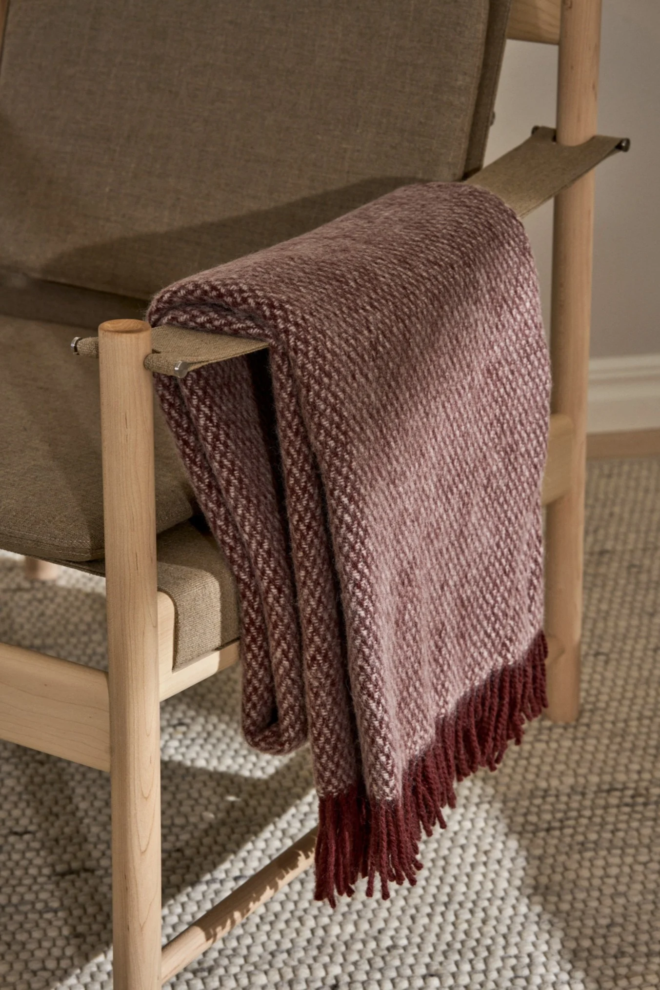 Reddish-brown patterned throw blanket with fringes draped over the arm of a light wood and olive green fabric chair on a patterned rug.