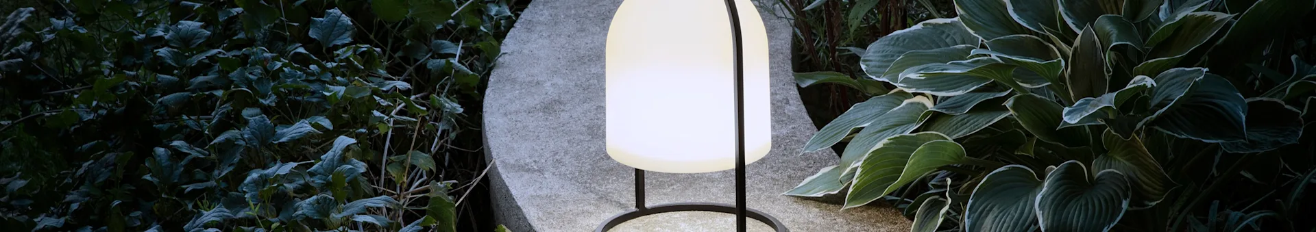 An illuminated outdoor lamp with a white oval shade and black frame sits on a stone base amidst dark green foliage.
